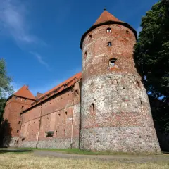 Zamek w Bytowie, potężna ceglana twierdza z kamienną podstawą wieży, otoczona zielenią pod błękitnym niebem.