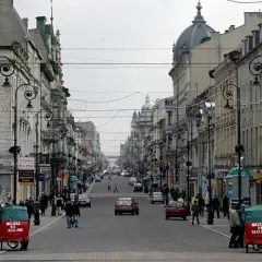 Ulica Piotrkowska w Łodzi, pełna zabytkowych kamienic, latarni i ruchu ulicznego. Dwa czerwone pojazdy z napisem "Miejsce na reklamę" stoją przy chodniku.