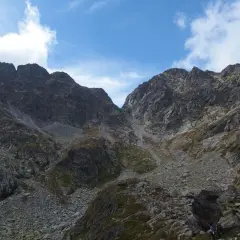 Kamienista dolina w górach, gdzie skalne zbocza tworzą wrażenie zawrotu głowy. W oddali widać turystę.