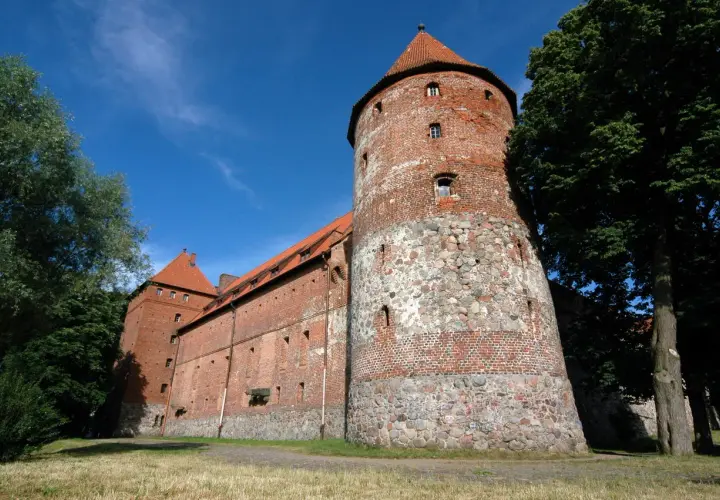 Zamek w Bytowie, potężna ceglana twierdza z kamienną podstawą wieży, otoczona zielenią pod błękitnym niebem.
