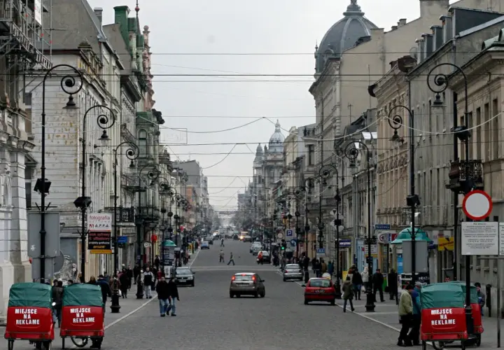 Ulica Piotrkowska w Łodzi, pełna zabytkowych kamienic, latarni i ruchu ulicznego. Dwa czerwone pojazdy z napisem "Miejsce na reklamę" stoją przy chodniku.
