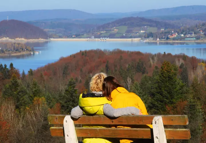 Para idealna na bieszczady na weekend. Dwie osoby siedzą na ławce, przytulając się, patrząc na jezioro i jesienne wzgórza.