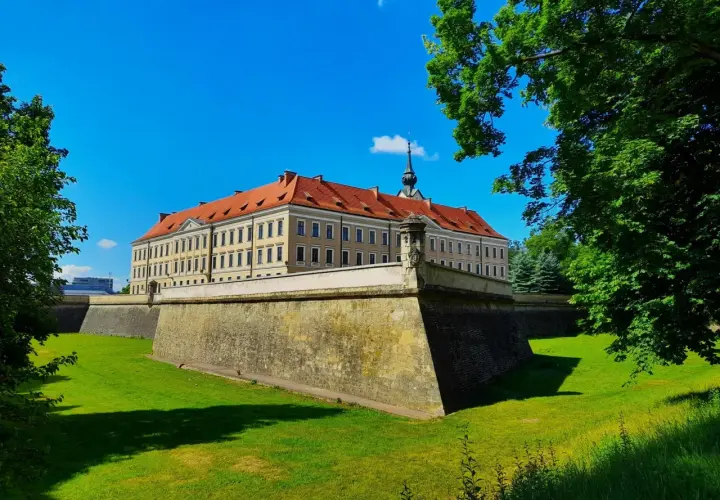 Zamek w Rzeszowie z czerwonym dachem i fosą otoczoną zielonym trawnikiem, pod błękitnym niebem.