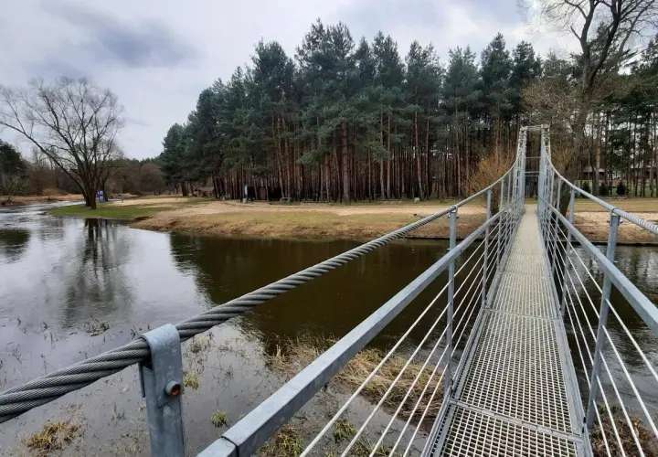 Metalowy most wiszący w Polsce nad rzeką, otoczony lasem sosnowym i piaszczystym brzegiem.