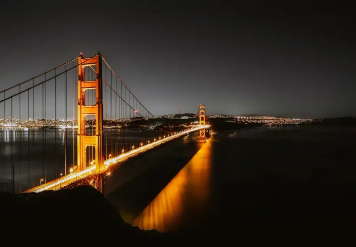 Złota Brama nocą, oświetlona, nad wodą. W oddali widać miasto w Kalifornii, San Francisco, z migoczącymi światłami.