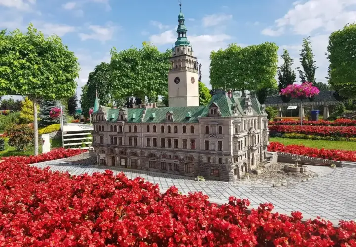 Miniatura ratusza z wieżą zegarową, otoczona czerwonymi kwiatami. Idealne miejsce na trasie po Kotlinie Kłodzkiej.