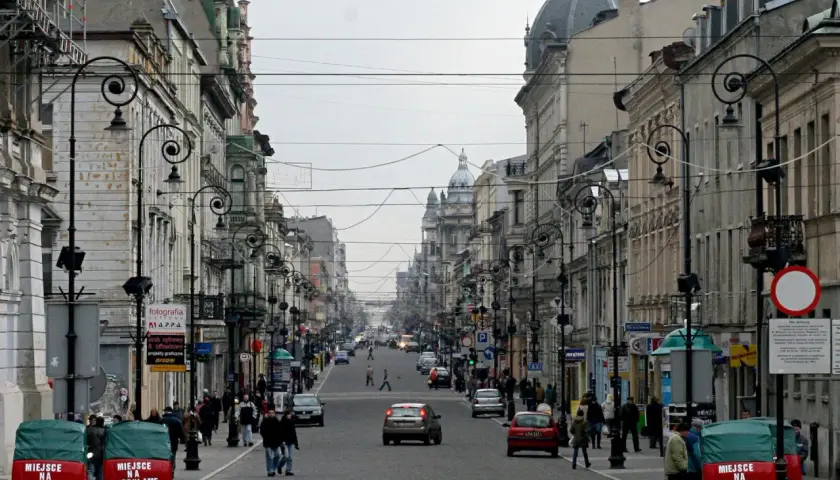 Ulica Piotrkowska w Łodzi, pełna zabytkowych kamienic, latarni i ruchu ulicznego. Dwa czerwone pojazdy z napisem "Miejsce na reklamę" stoją przy chodniku.