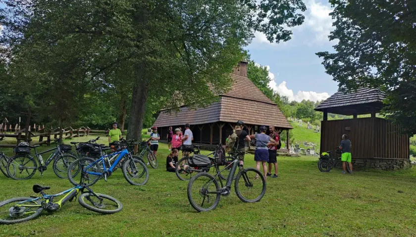 Grupa rowerzystów odpoczywa przy drewnianej cerkwi, planując dalszą podróż po malowniczych trasach rowerowych Bieszczad.
