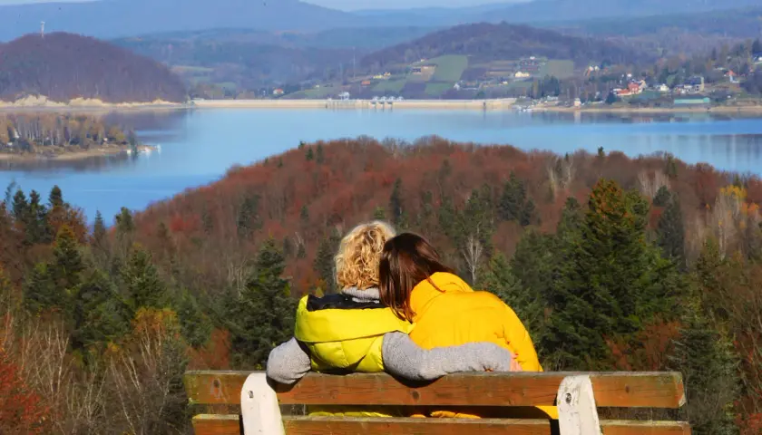 Para idealna na bieszczady na weekend. Dwie osoby siedzą na ławce, przytulając się, patrząc na jezioro i jesienne wzgórza.