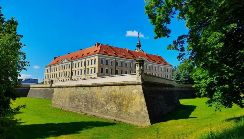 Zamek w Rzeszowie z czerwonym dachem i fosą otoczoną zielonym trawnikiem, pod błękitnym niebem.