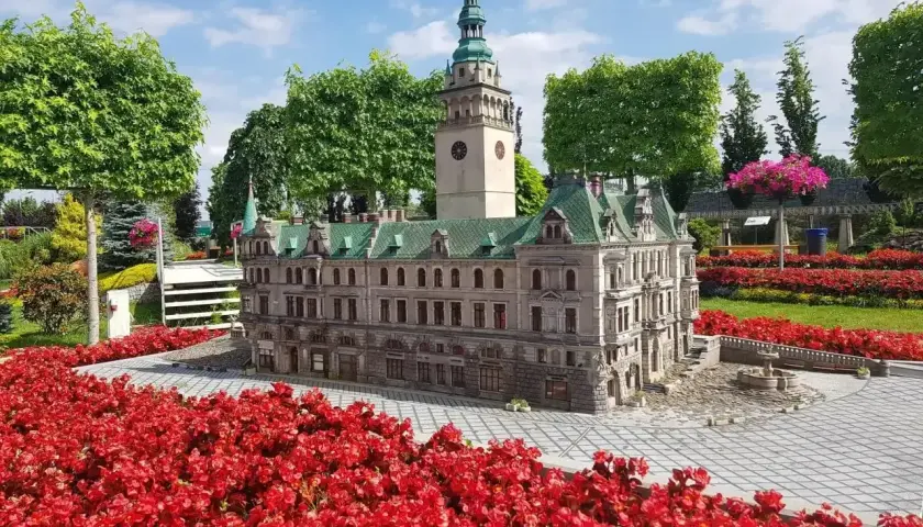 Miniatura ratusza z wieżą zegarową, otoczona czerwonymi kwiatami. Idealne miejsce na trasie po Kotlinie Kłodzkiej.