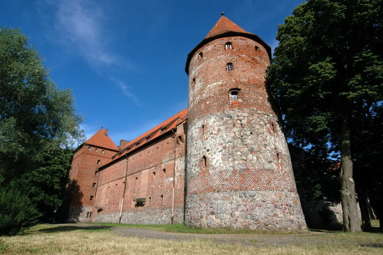 Zamek w Bytowie, potężna ceglana twierdza z kamienną podstawą wieży, otoczona zielenią pod błękitnym niebem.