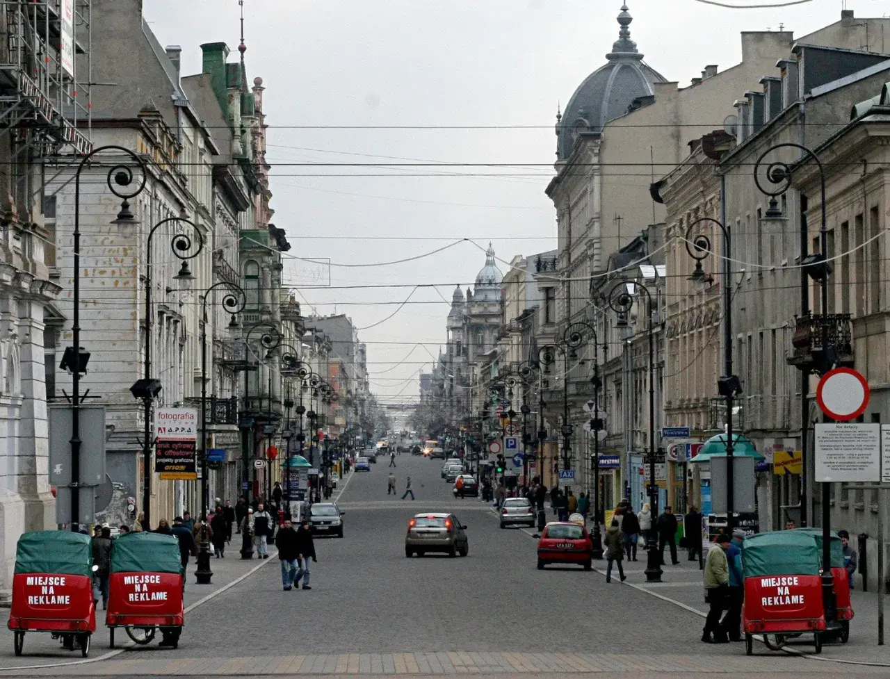Ulica Piotrkowska w Łodzi, pełna zabytkowych kamienic, latarni i ruchu ulicznego. Dwa czerwone pojazdy z napisem "Miejsce na reklamę" stoją przy chodniku.
