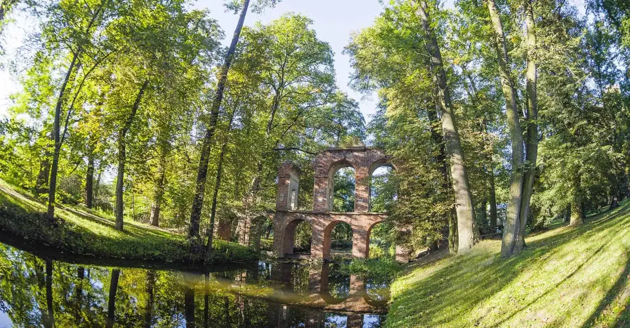Ruiny mostu z cegły w parku, otoczone drzewami. Woda odbija zieleń i architekturę, tworząc malowniczy widok na pałac w Nieborowie.