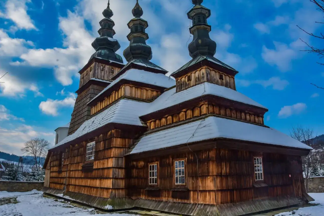Drewniana cerkiew w zimowej scenerii Beskidu Niskiego, z kopułami i ośnieżonymi dachami, pod błękitnym niebem.